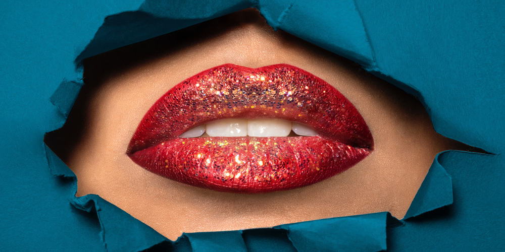 A womens mouth with red lipstick and glitter slightly opeing her mouth, showing through a hole in a blue piece of paper