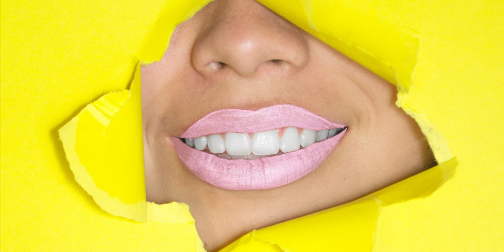 A womens bottom part of her face showing through a ripped hole in a yellow piece of paper, women has pink lipstick and white porcelain veneers